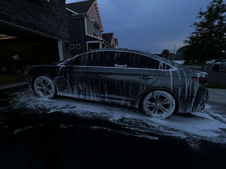 Dark sedan covered in soap suds during twilight car wash in residential driveway
